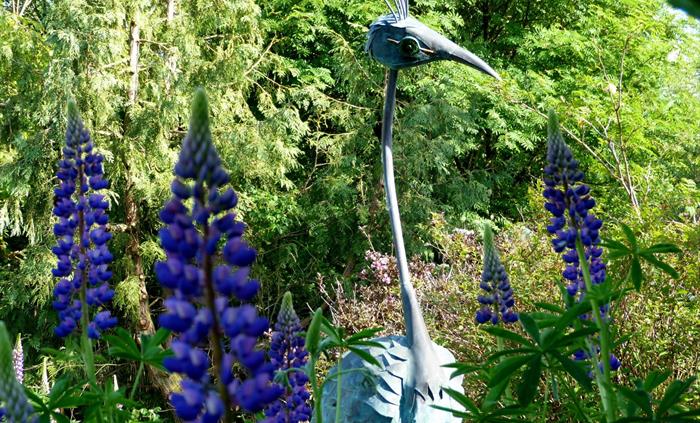 Sculpture of a fantasy peacock made of verdigris copper and glass. Blue lupins in the foreground
